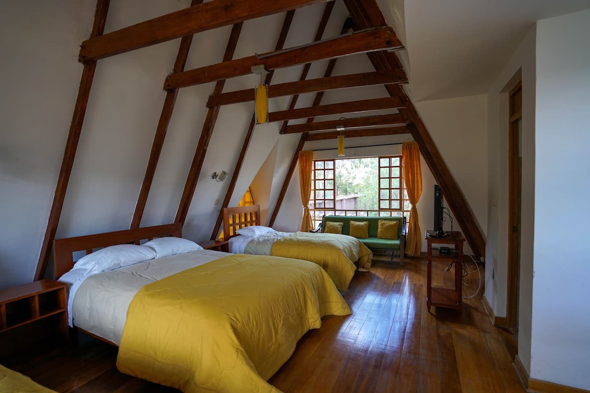 Una habitación con techo inclinado, vigas de madera, dos camas con ropa de cama amarilla, un sofá verde y una ventana con cortinas.