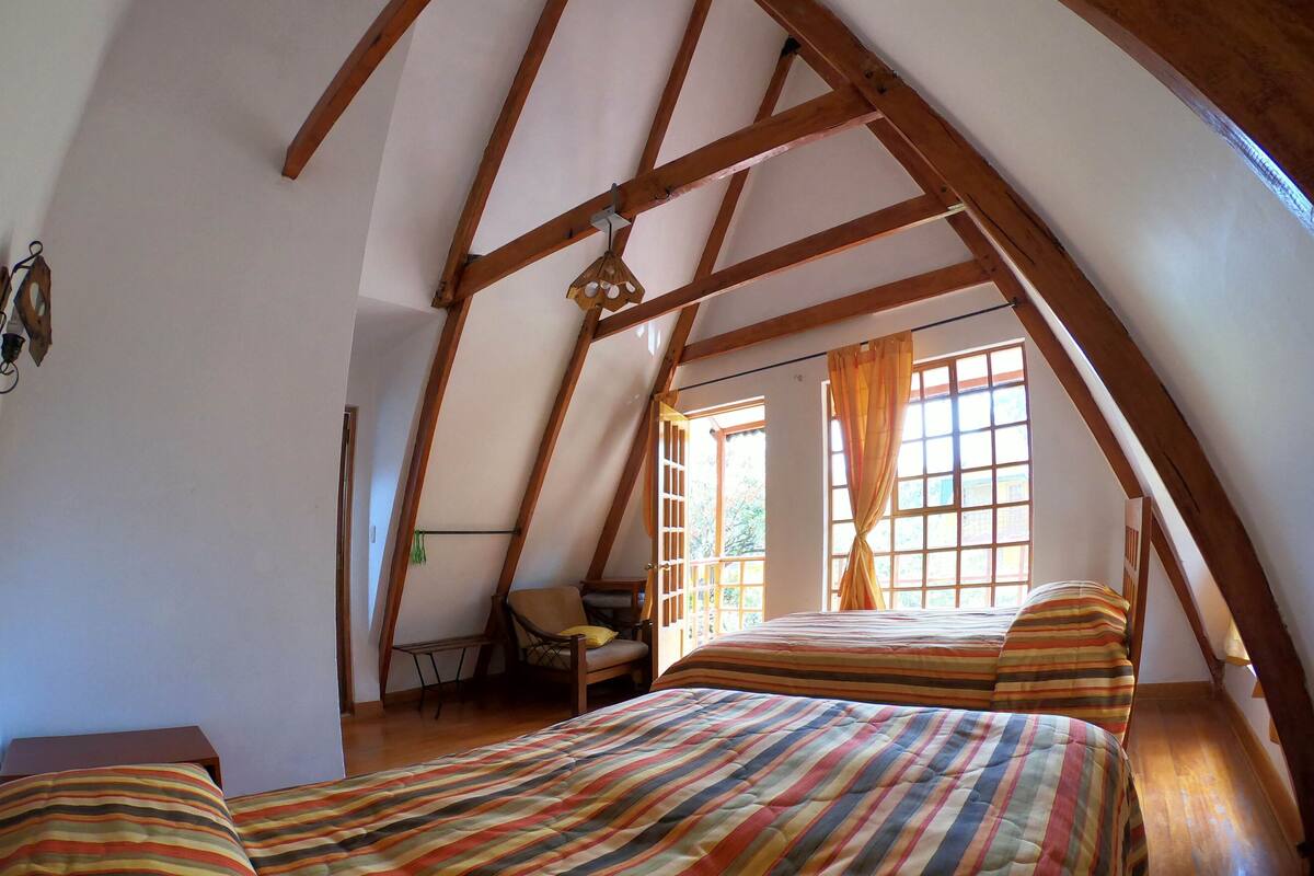 Un dormitorio con techo inclinado, vigas de madera, cama con ropa de rayas, una silla y un ventanal con cortinas.