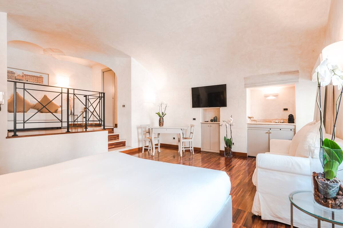 A modern hotel room with a large bed, a dining area, a kitchen, and a TV mounted on the wall.