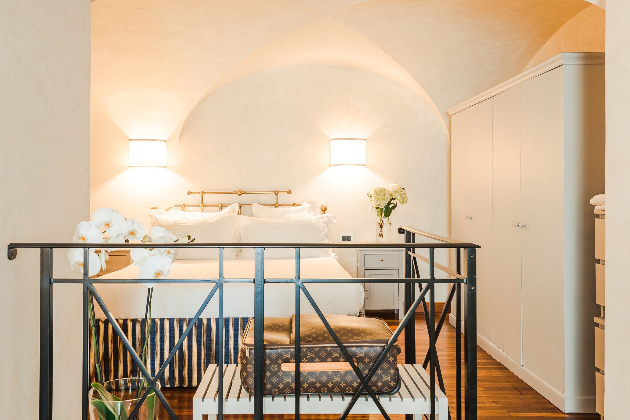 A bedroom with a bed, bedside lamps, a nightstand, a suitcase, and a view of a hallway.