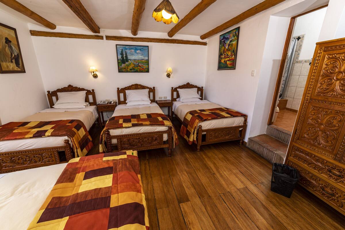 A room with three wooden beds, each with a quilt. There are wall-mounted lights and paintings on the walls.