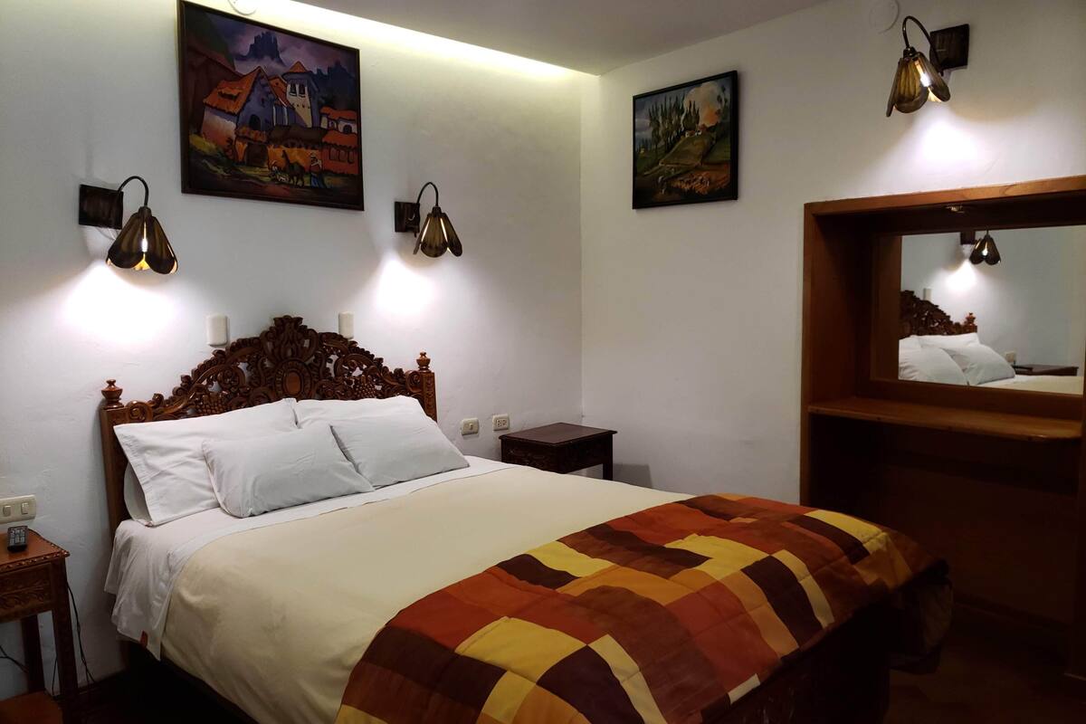 A neatly made bed with a patterned blanket, two wall-mounted lamps, and two paintings on the wall.