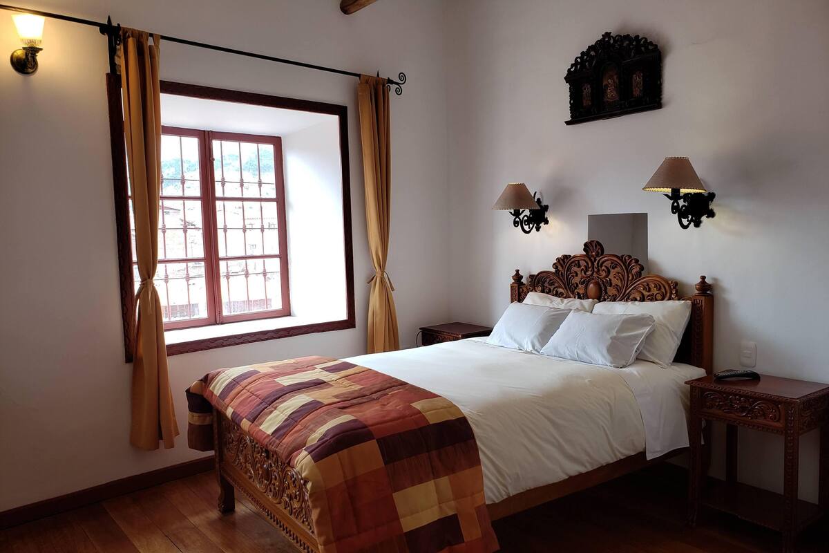 A bedroom with a wooden bed, a window with curtains, a nightstand, and wall-mounted lights.