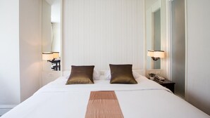 Ein Hotelzimmer mit einem großen Bett, zwei Kissen, einem Nachttisch mit Lampe, einem Spiegel und einem an der Wand befestigten Telefon.