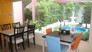 A dining area with a table and chairs, a small fountain, and a view of a garden with a white fence and greenery. - C2 Residence Boutique Hotel (Lampang)