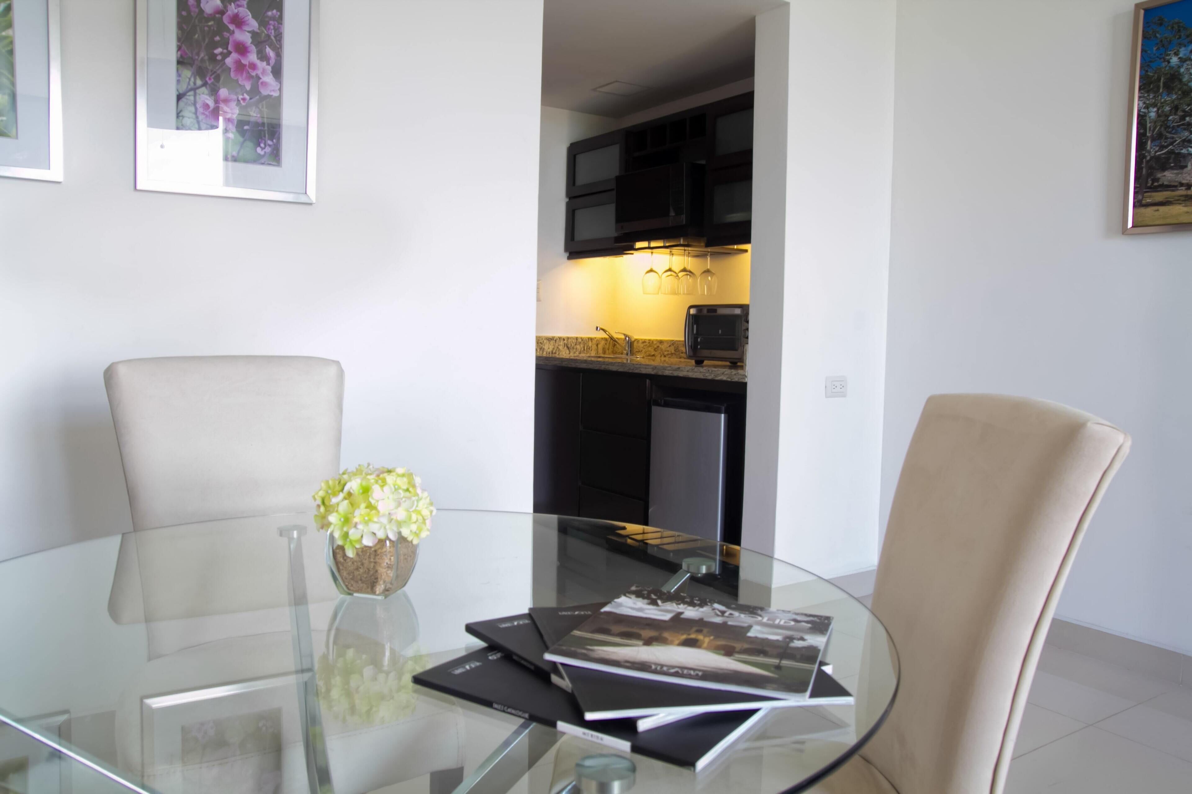 A modern dining area with a glass table, two chairs, a vase with flowers, and framed pictures on the wall.