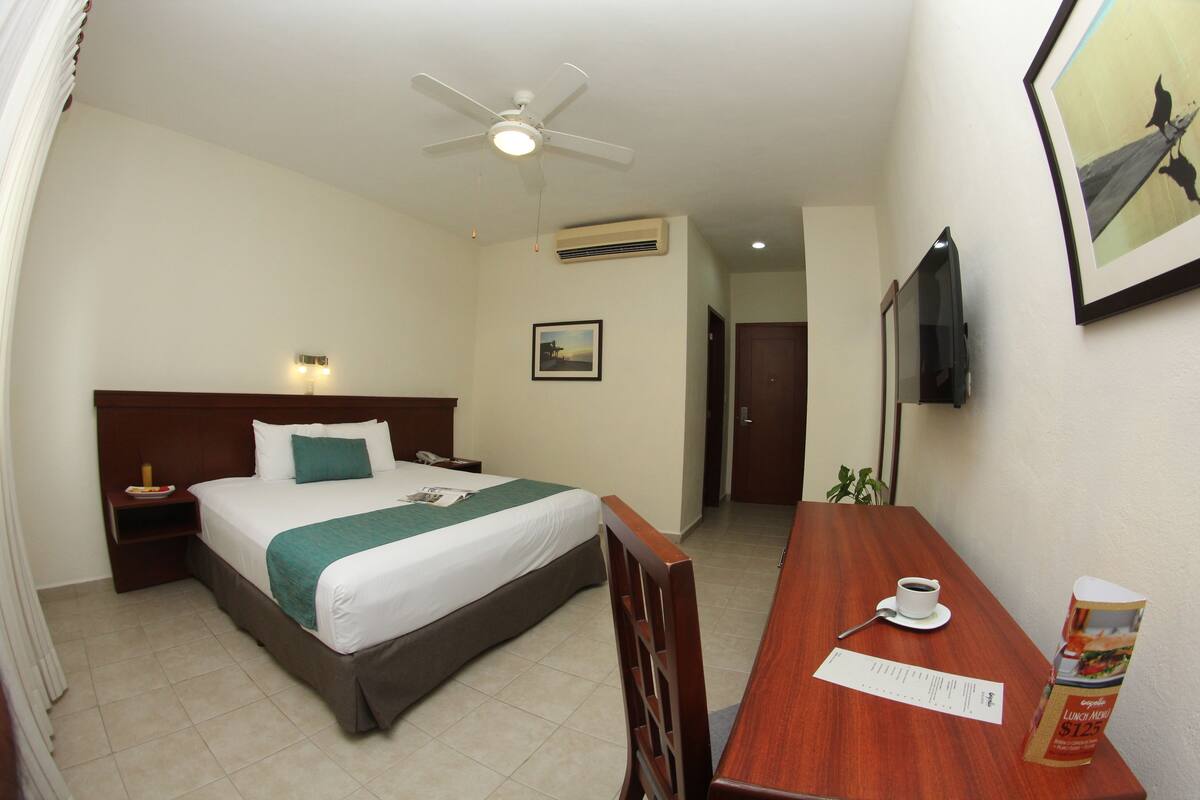 Una habitación de hotel con cama, una mesa de madera con un menú, un ventilador de techo, un equipo de aire acondicionado y un televisor en la pared.