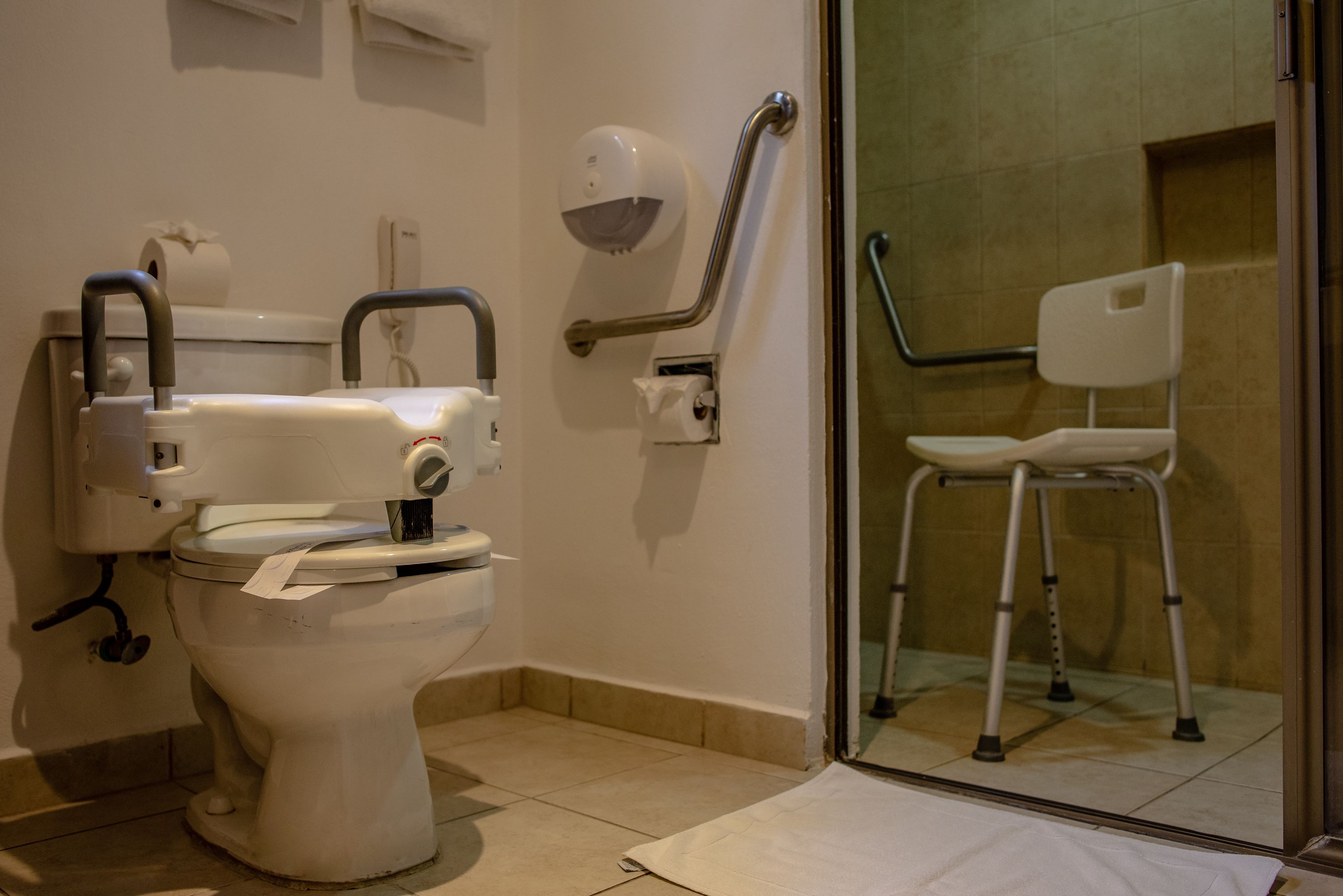 A bathroom with a white toilet, a handrail, a white chair, and a glass shower enclosure.