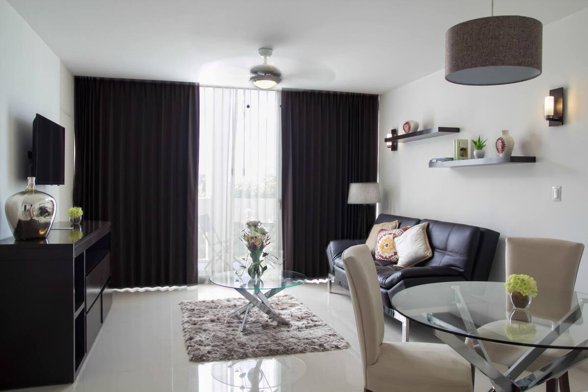 Una sala de estar moderna con una mesa de centro de vidrio, un sofá, una silla, un televisor y un ventilador de techo.
