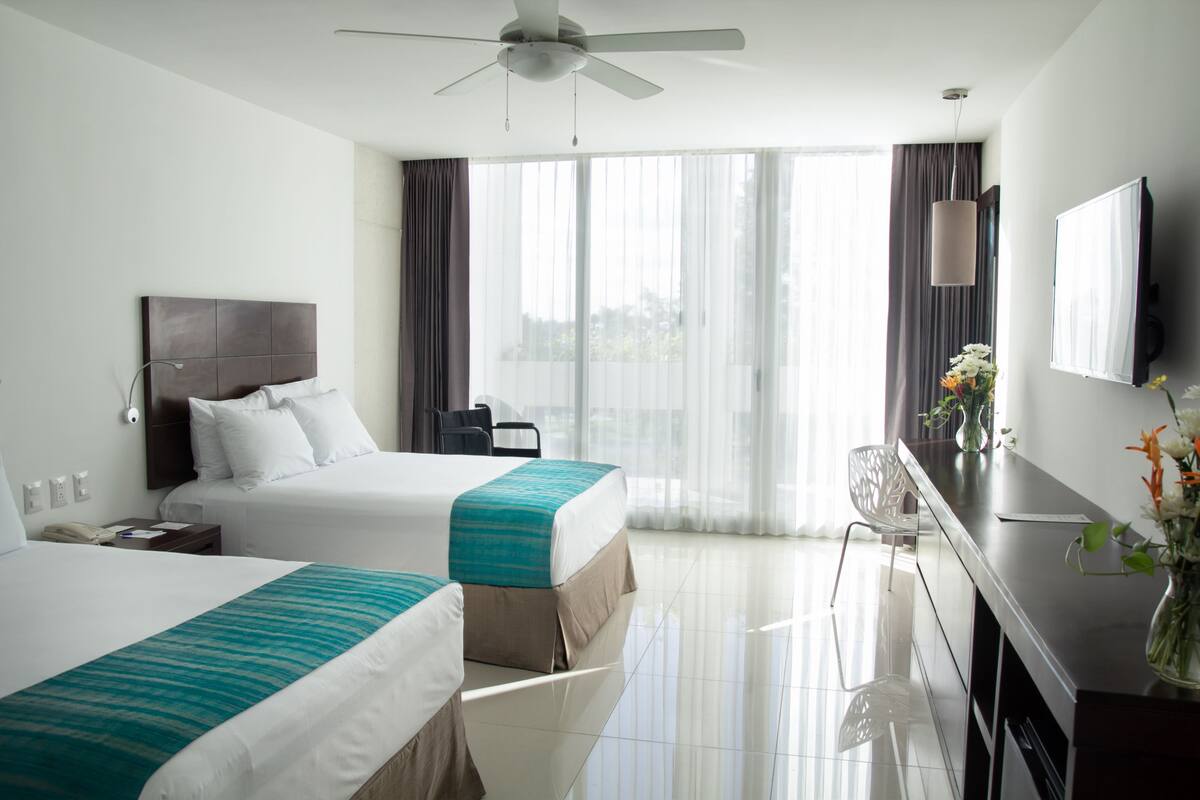 Habitación de hotel con dos camas, un escritorio, una silla, un televisor y un ventanal con cortinas.