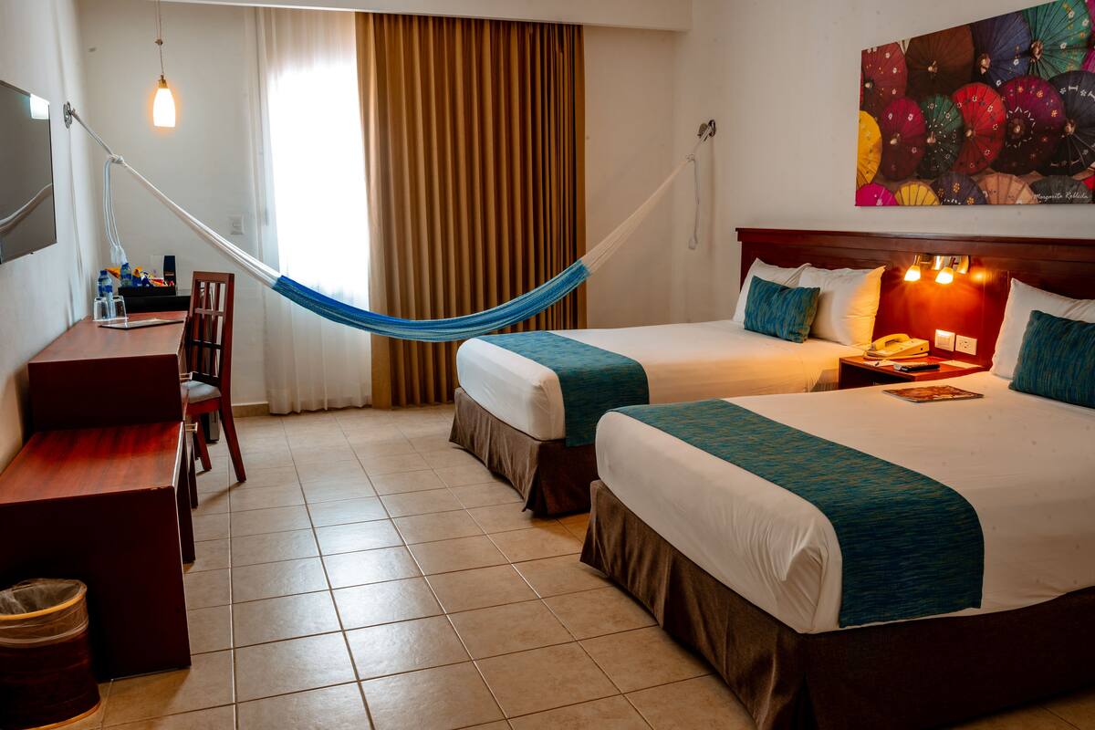 Habitación de hotel con dos camas, un escritorio, una silla, una lámpara de escritorio, una hamaca y una obra de arte con un paraguas colorido en la pared.