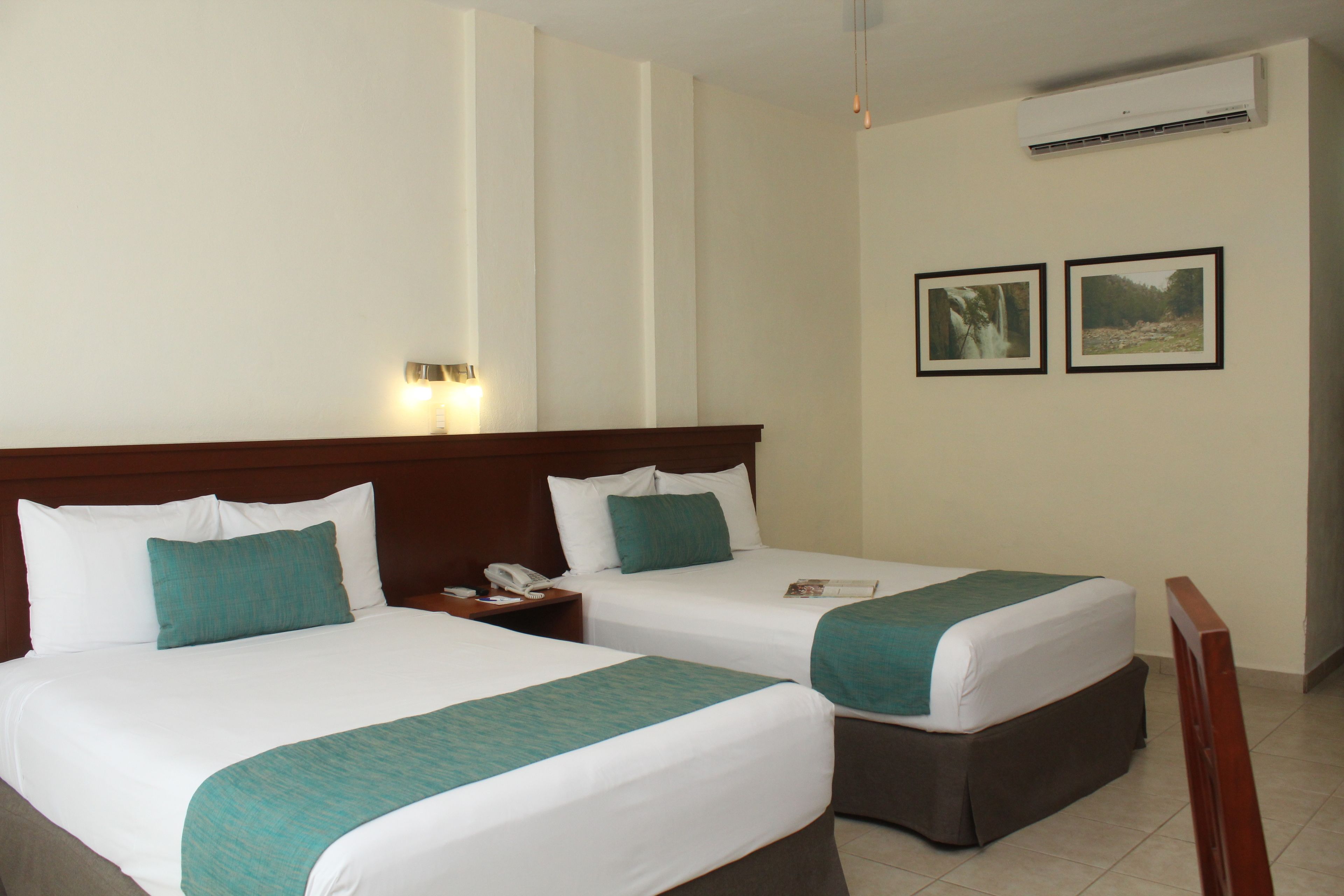 A hotel room with two beds, each with white linens and teal accents, a wooden headboard, a bedside table with a lamp, and two framed pictures on the wall.