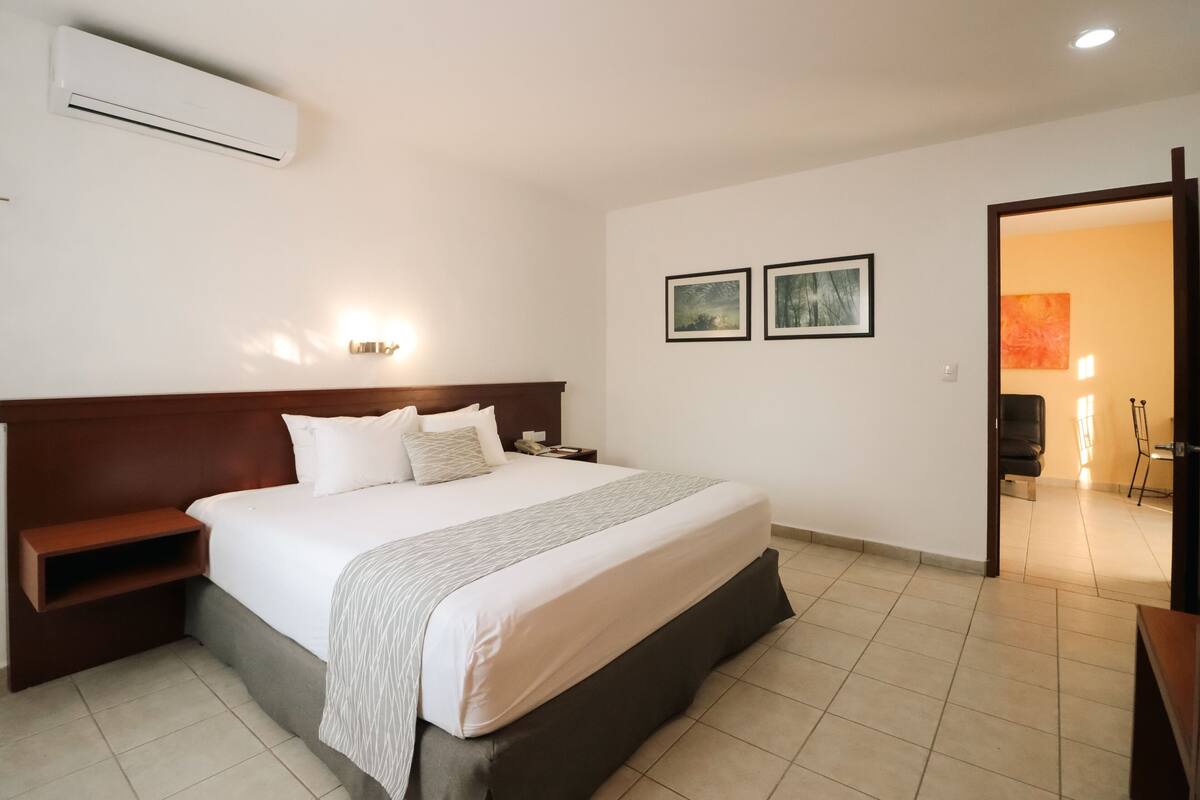 Habitación de hotel con una cama grande, una mesita de noche, un aire acondicionado de pared y dos cuadros enmarcados.