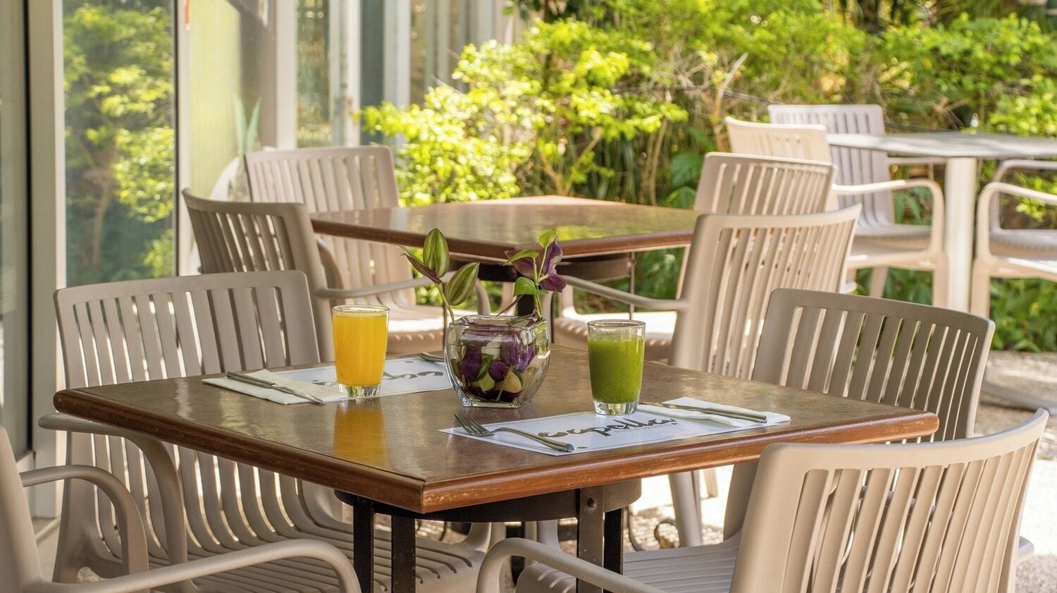 Una mesa con dos bebidas: un vaso de jugo de naranja y un vaso de jugo verde, además de un pequeño florero con flores.