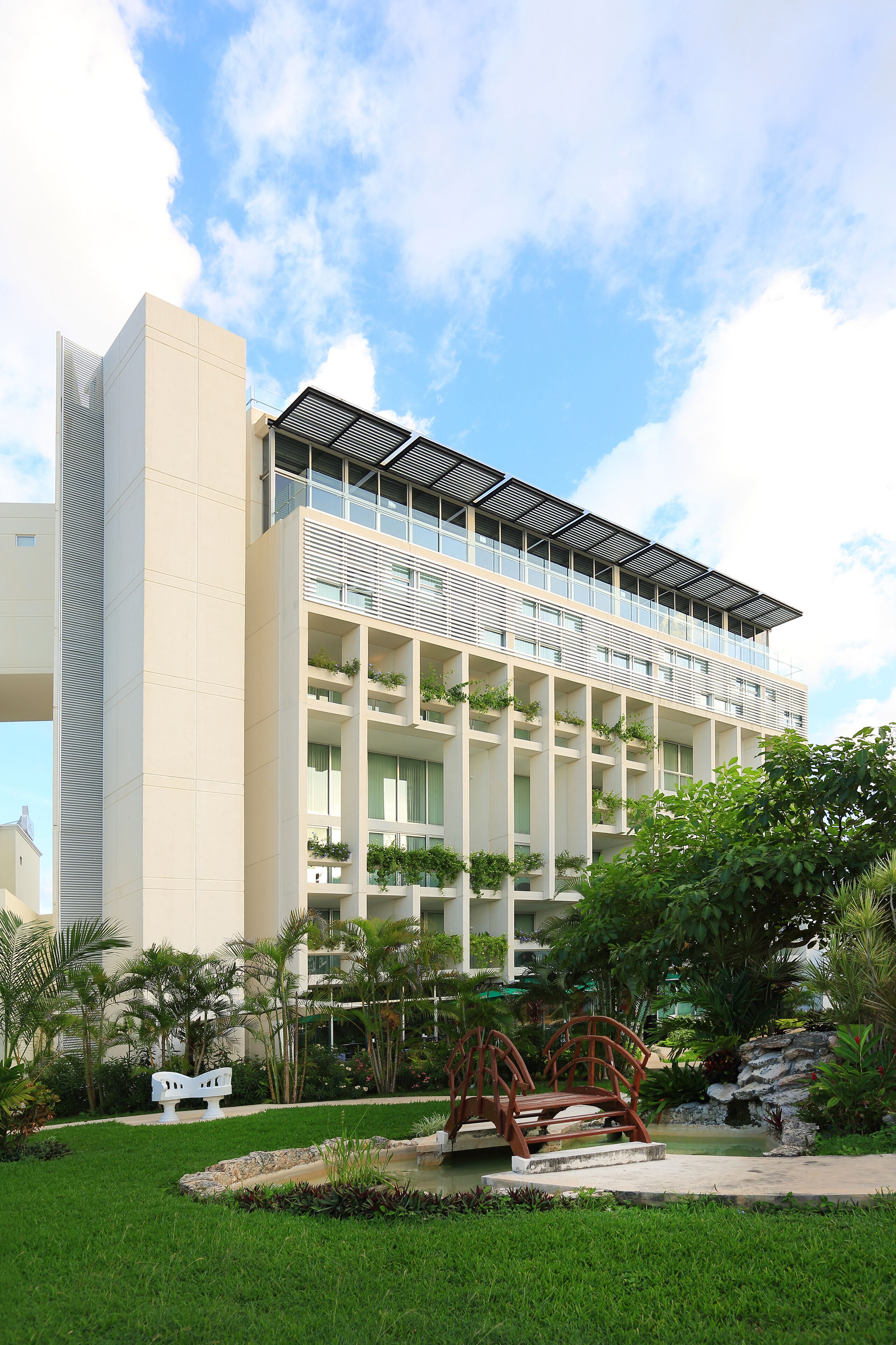 Un hotel moderno con techo plano, varios balcones y un jardín con un puente y asientos.