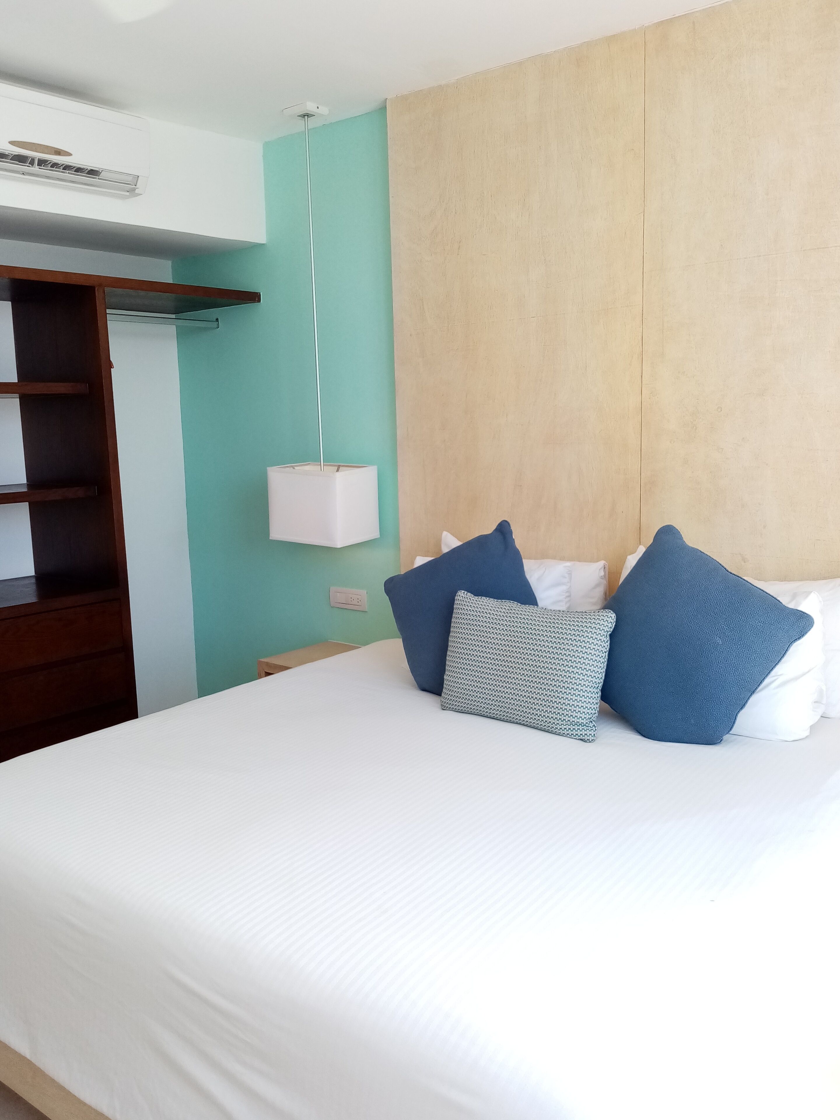 Cama bem arrumada com roupa de cama branca e almofadas azuis e estampadas, cabeceira de madeira, abajur branco e guarda-roupa de madeira.