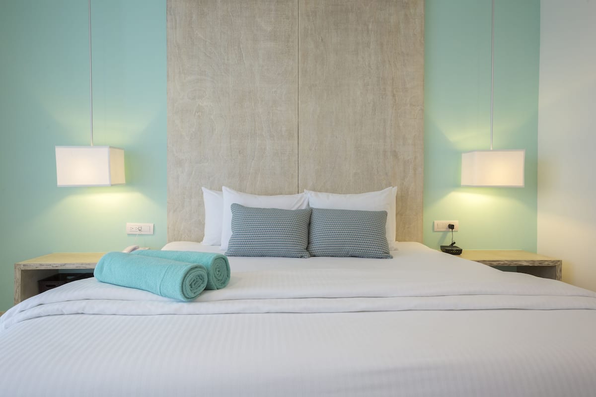 Una cama bien tendida con sábanas blancas y dos almohadas azules, una toalla verde aguamarina y dos lámparas de noche sobre mesitas de madera.