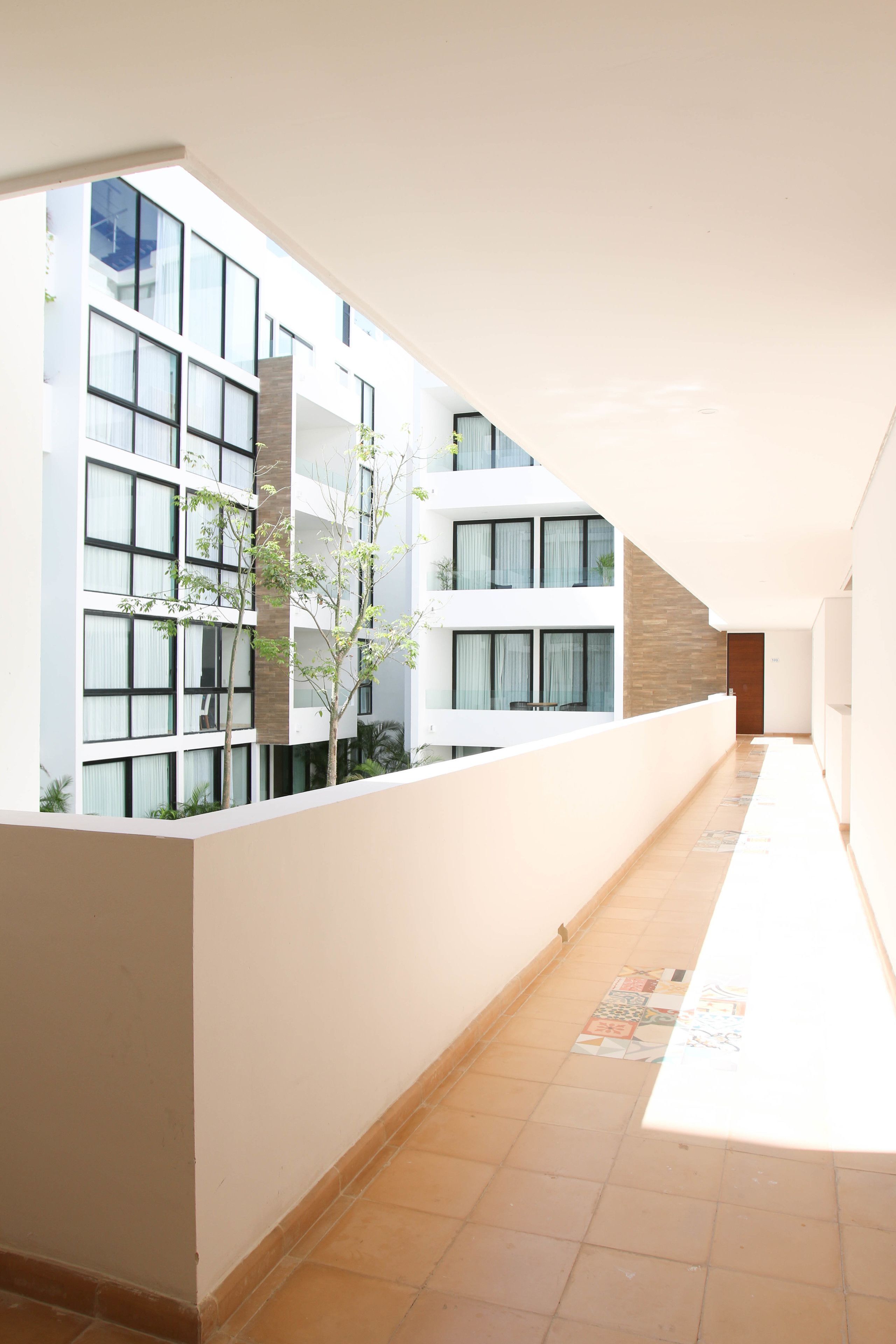 Un pasillo con piso de baldosas, paredes blancas y un edificio moderno con ventanas amplias y un árbol.