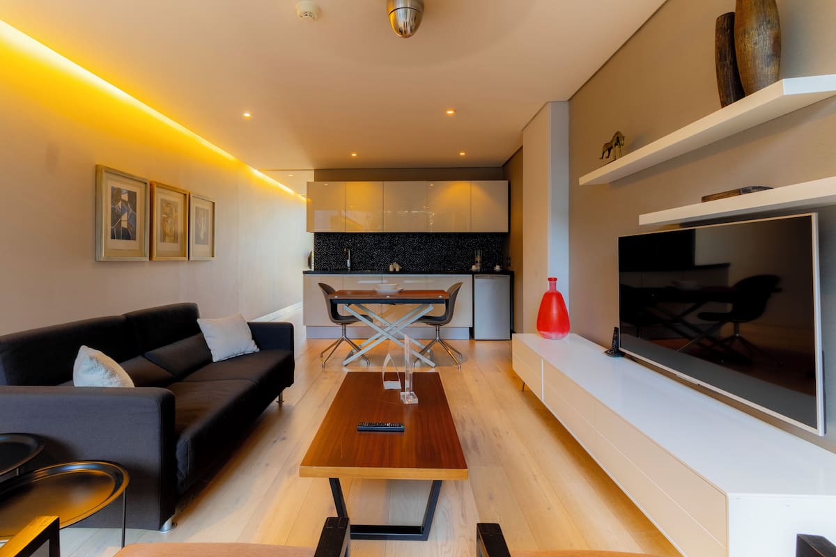 A modern living room with a sofa, coffee table, and TV. There is a kitchen in the background with a dining area.