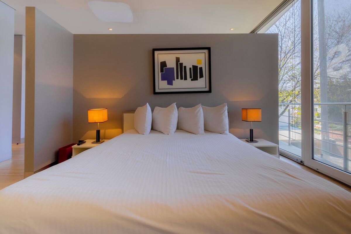 A modern hotel room with a large bed, two bedside lamps, a framed artwork on the wall, and a view through the window.