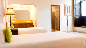 Una habitación de hotel con una cama, una mesita de noche, una lámpara, un espejo, un baño con un lavabo y una puerta que da a otra habitación.
