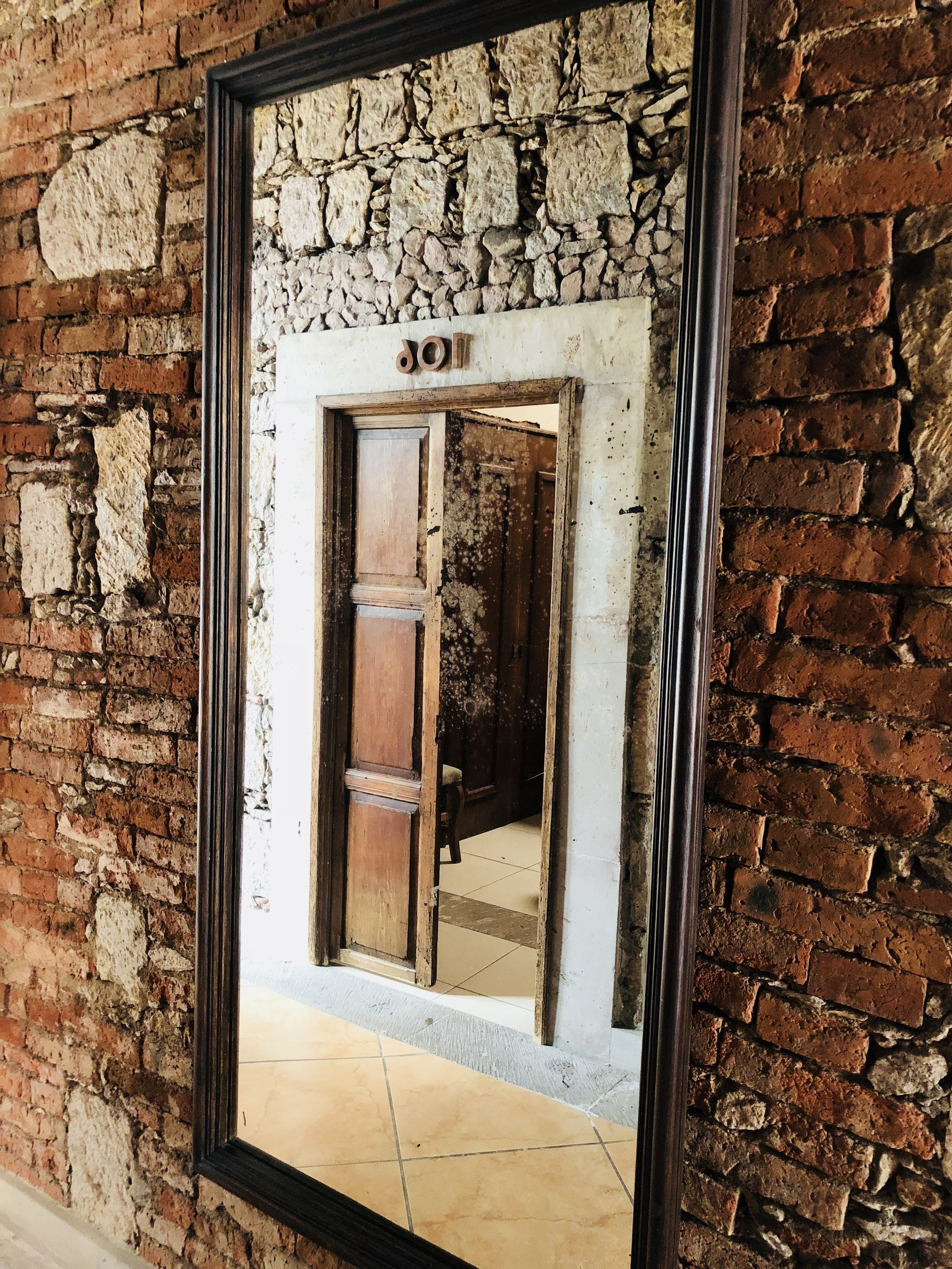 Un espejo refleja una habitación con pared de piedra, puerta de madera y suelo de baldosas.