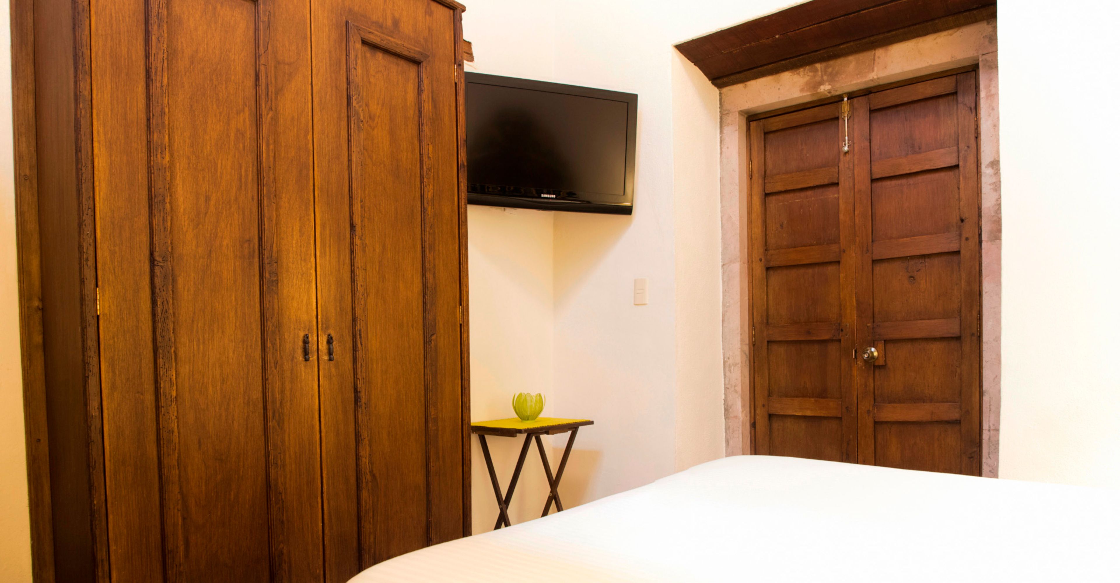 A bedroom with a bed, wooden wardrobe, a small table with a green object, and a TV mounted on the wall.