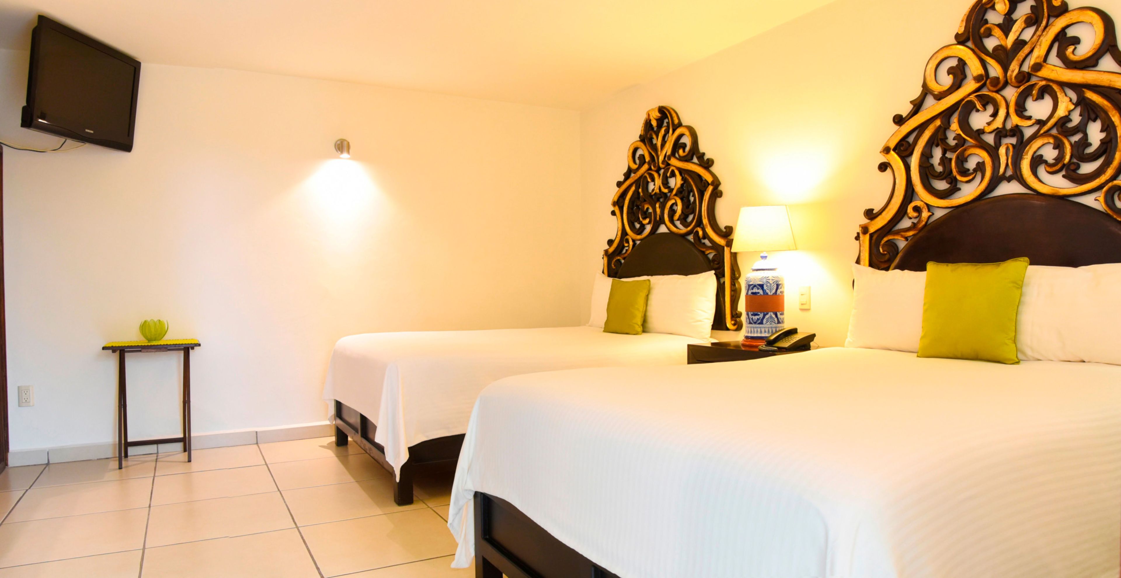 A hotel room with two beds, a small table, a lamp, and a television on the wall.