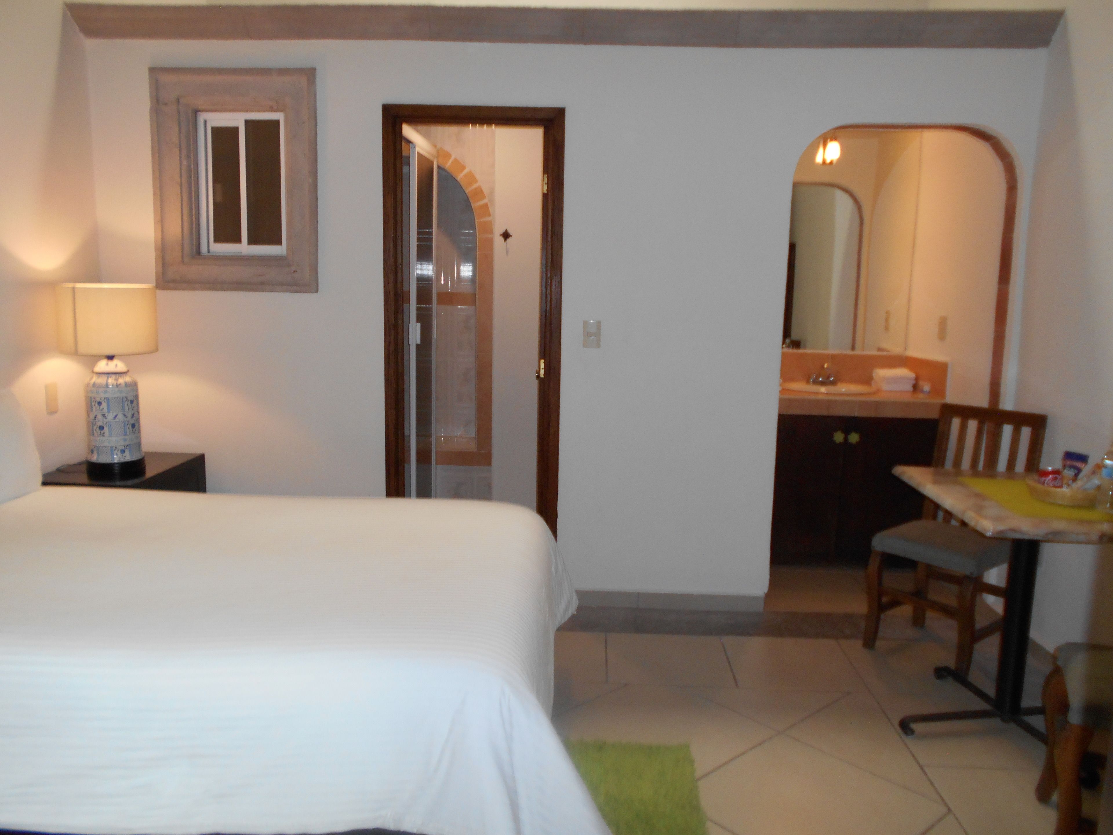 A hotel room with a bed, a nightstand with a lamp, a bathroom with a mirror, and a small table with a chair.