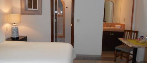 A hotel room with a bed, a nightstand with a lamp, a bathroom with a mirror, and a small table with a chair.
