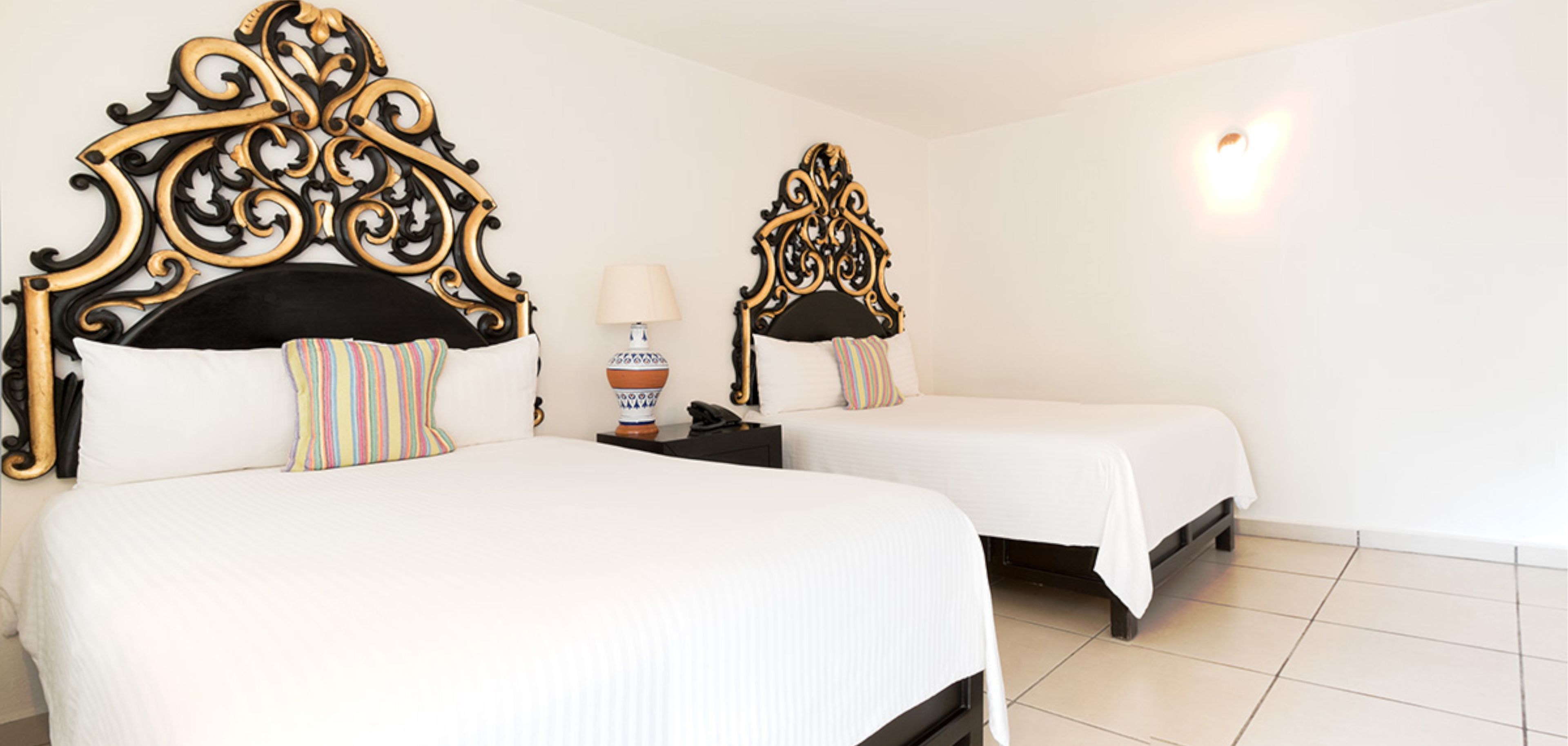 Two beds with ornate headboards, white bedding, and a bedside table with a lamp and a decorative vase.