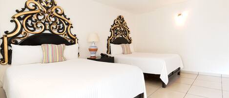 Two beds with ornate headboards, white bedding, and a bedside table with a lamp and a decorative vase.