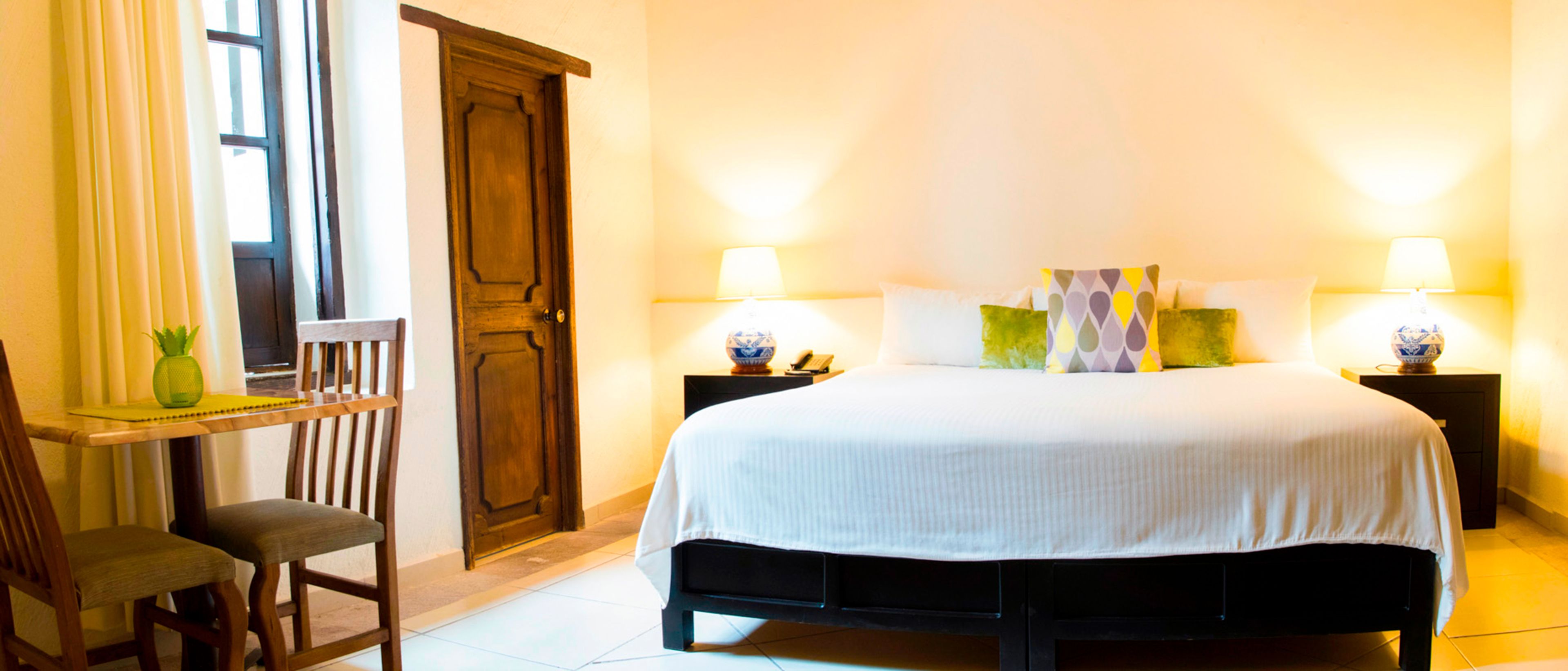 Une chambre d’hôtel avec un grand lit, deux tables de chevet avec des lampes, une chaise en bois, un bureau et une porte.