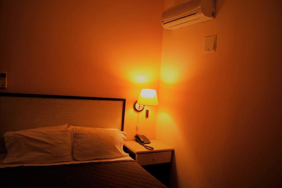 A hotel room with a bed, a bedside table with a lamp, and an air conditioner mounted on the wall.