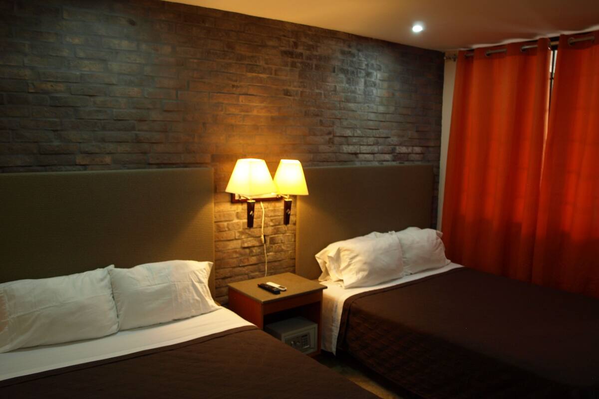A hotel room with two beds, a brick wall, and a nightstand with lamps.