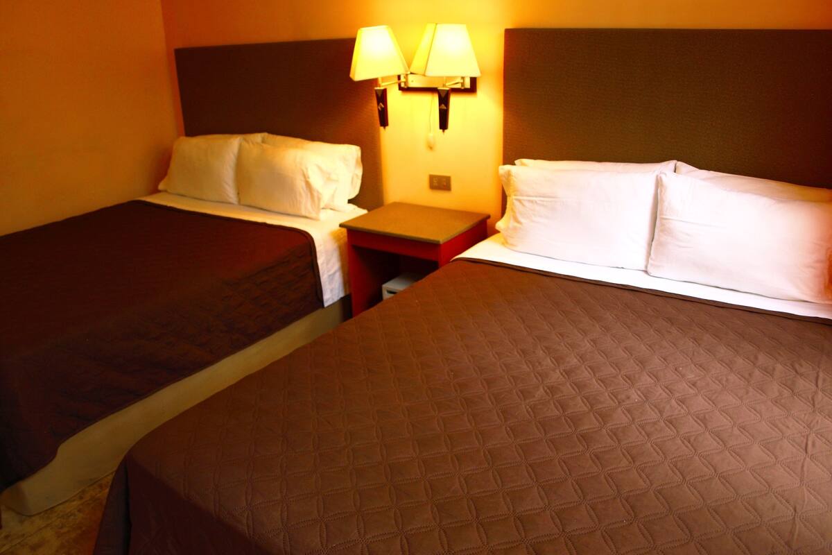 A hotel room with two beds, each with white linens, a nightstand with a lamp, and a headboard.