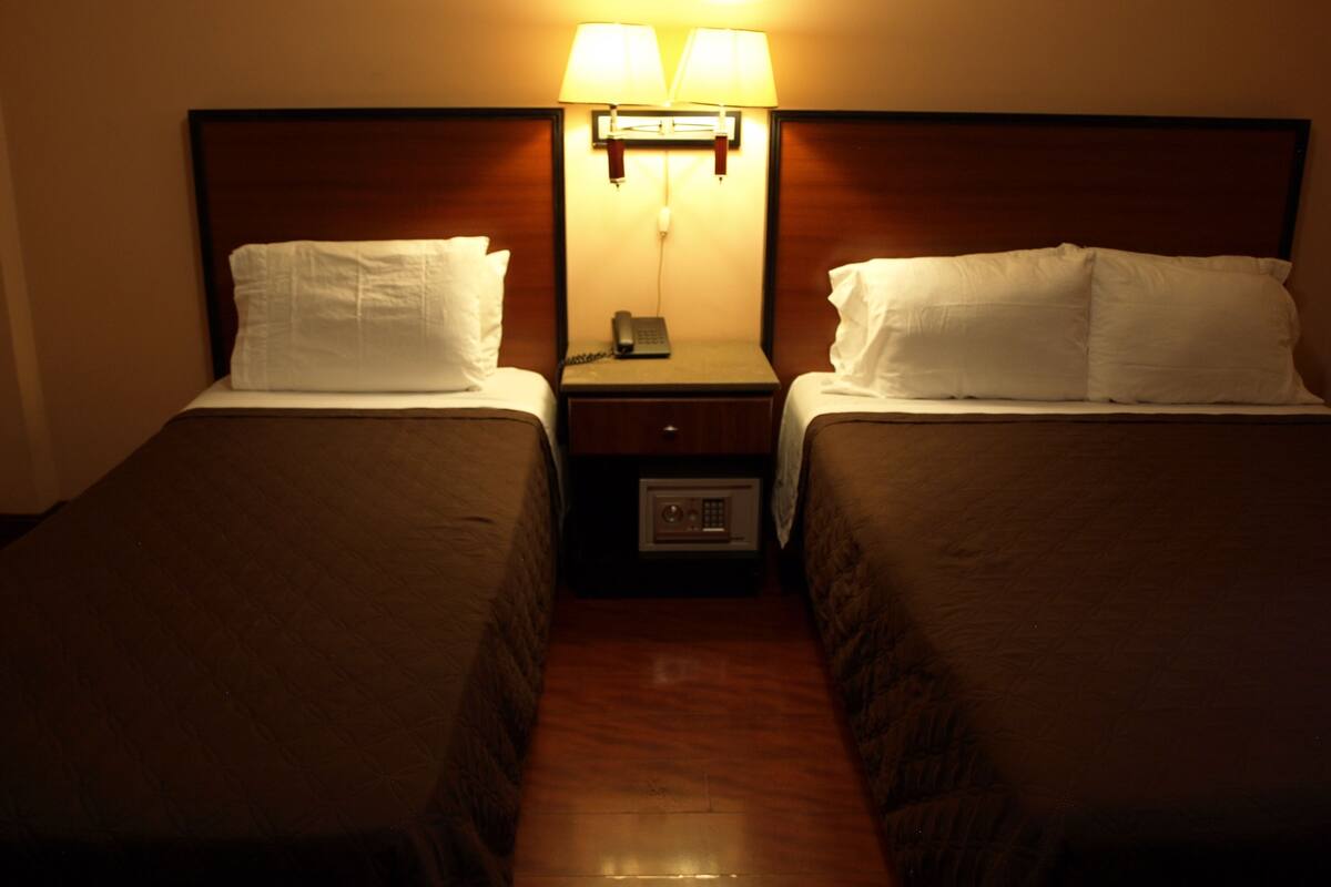A hotel room with two single beds, each with a pillow and a headboard. There is a nightstand between the beds with a lamp and a phone.