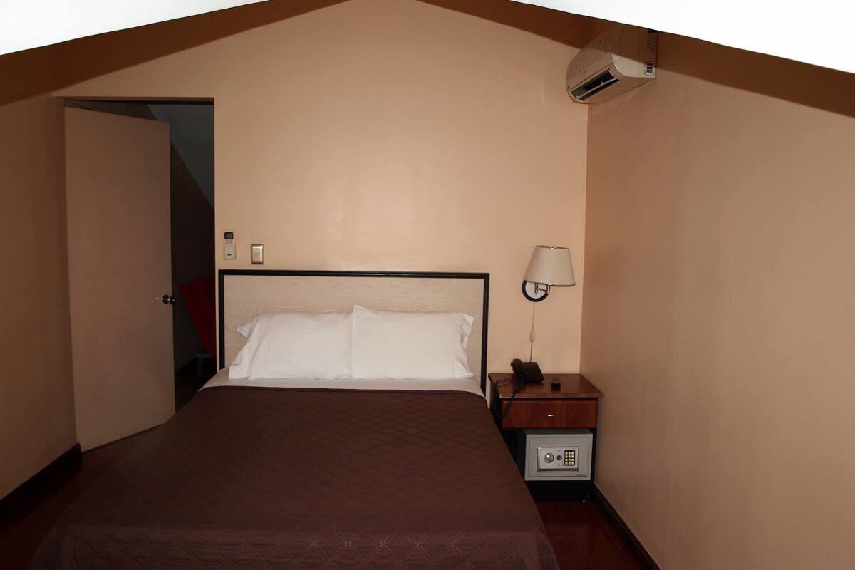A hotel room with a bed, bedside lamp, nightstand, and a safe.