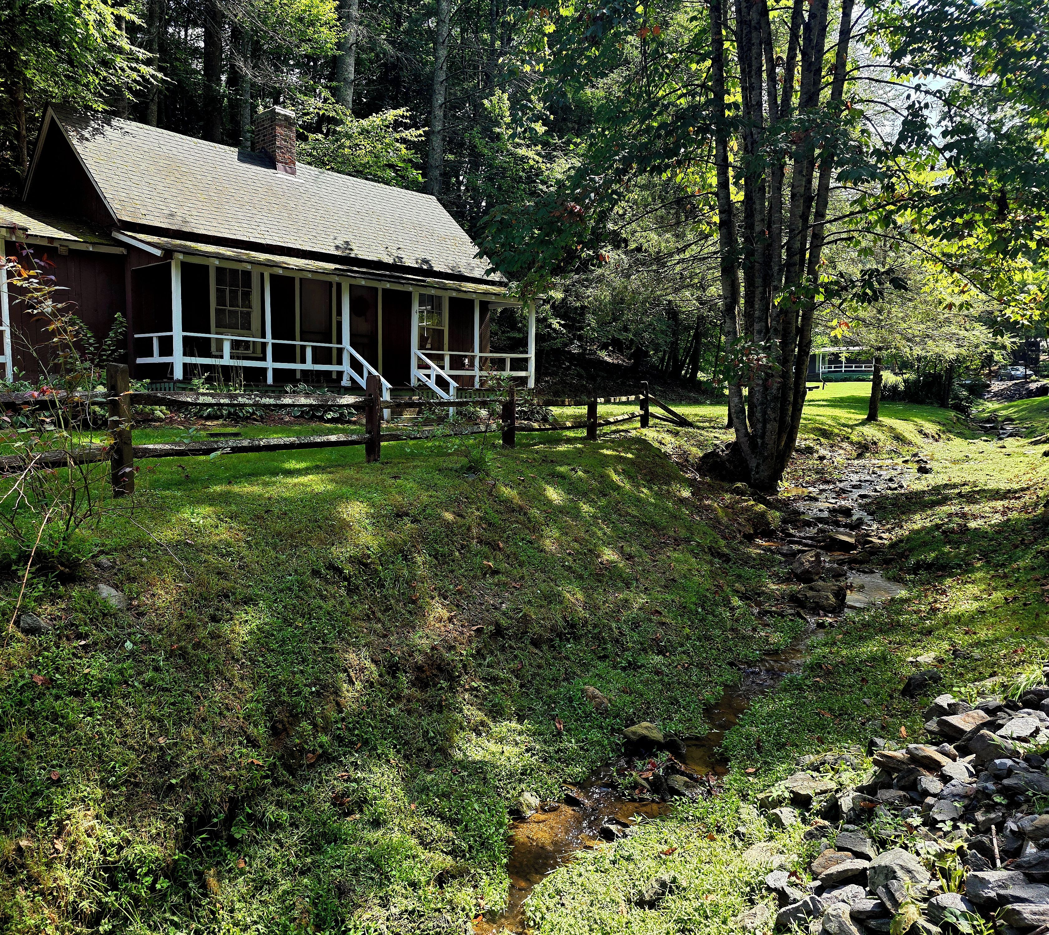 A rustic cabin with a porch, surrounded by dense forest and a small stream.