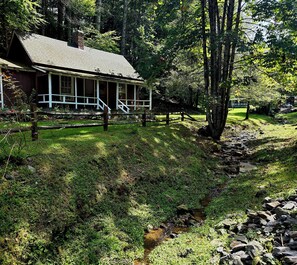 Una cabaña rústica con un porche, rodeada de un bosque denso y un pequeño arroyo.
