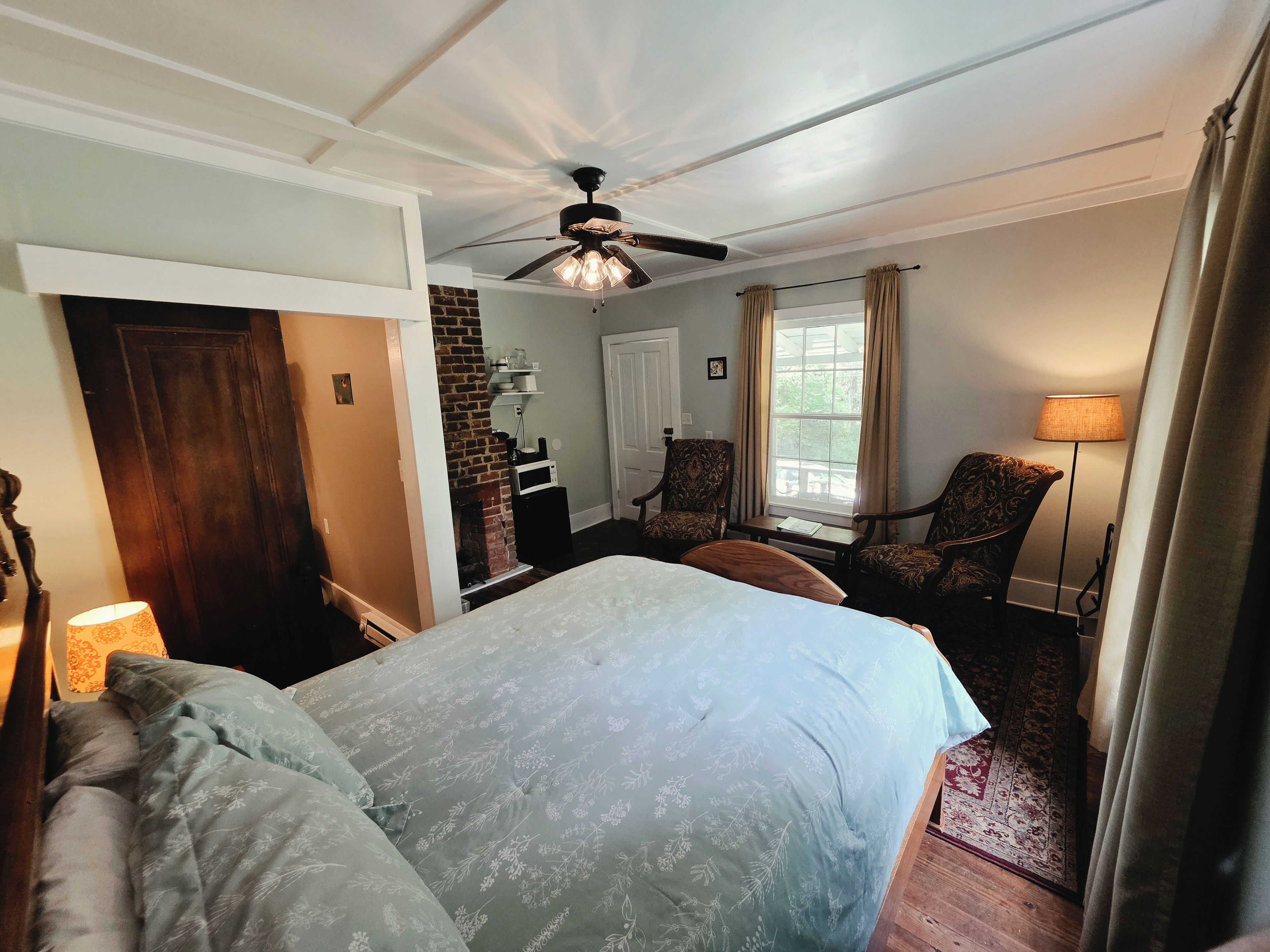 A hotel room with a bed, a fireplace, a ceiling fan, a window with curtains, and a lamp.