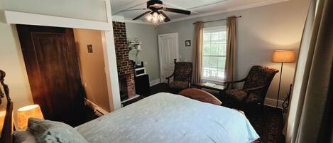 A hotel room with a bed, a fireplace, a ceiling fan, a window with curtains, and a lamp.