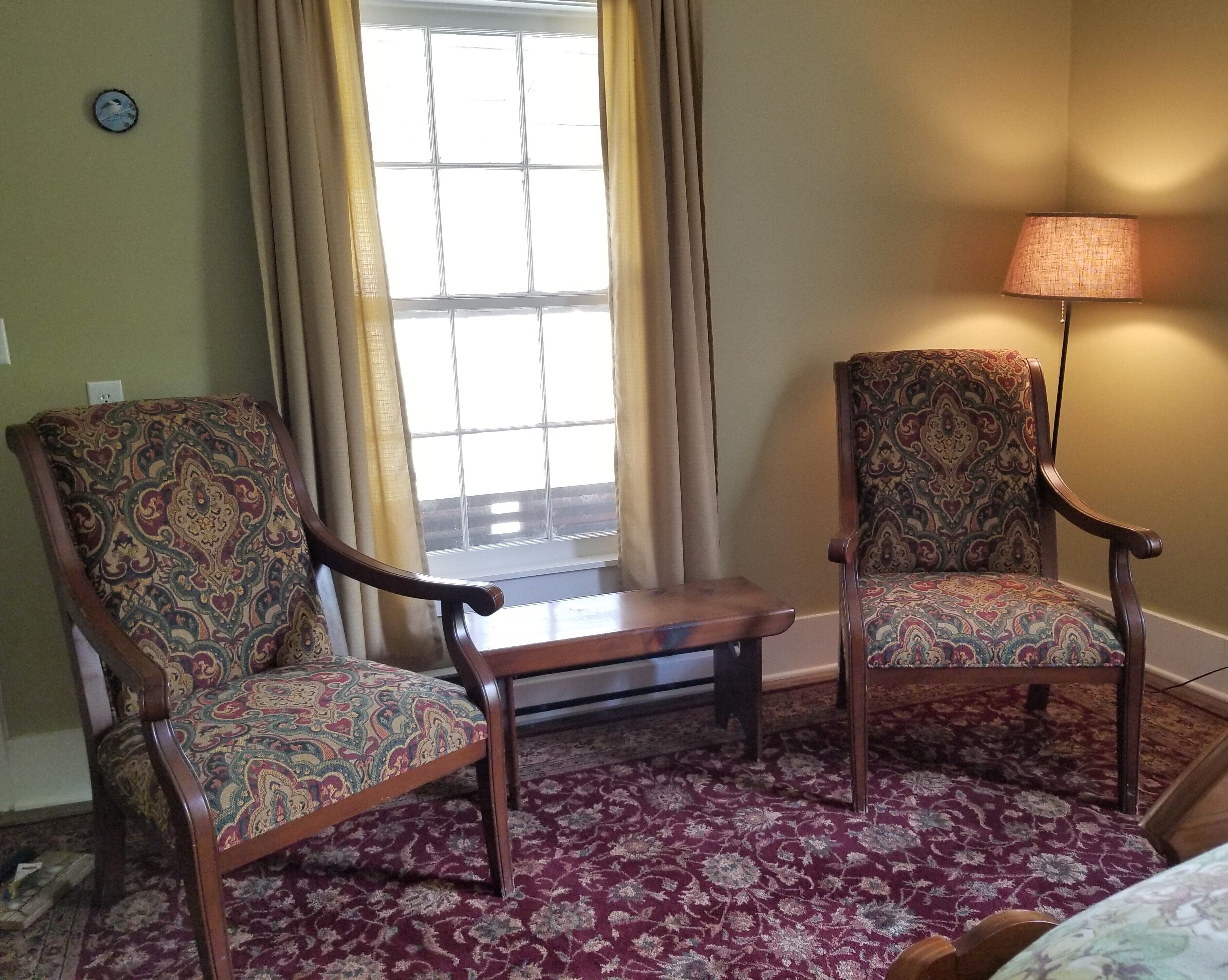 A room with two patterned armchairs, a small table, a lamp, and a window with curtains.