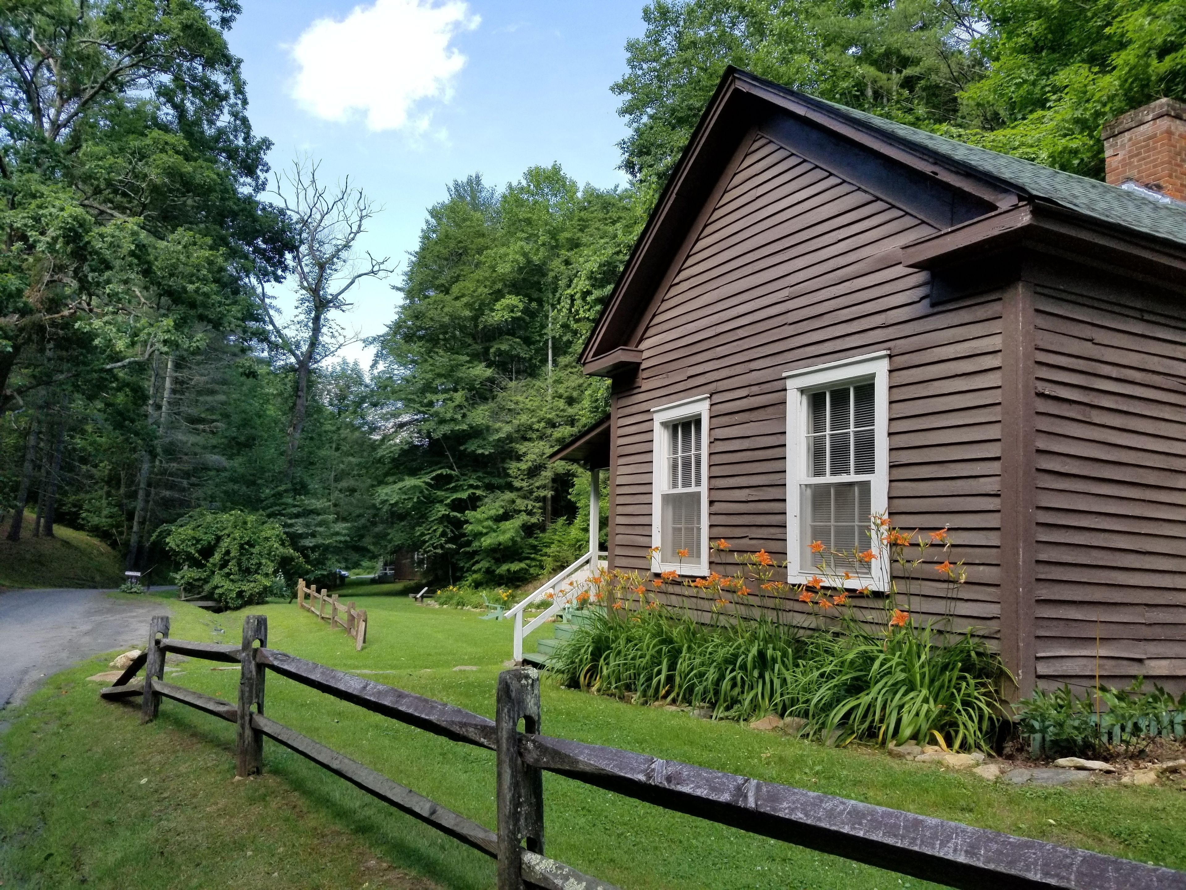 A wooden cabin with a sloped roof, surrounded by lush greenery and a wooden fence.