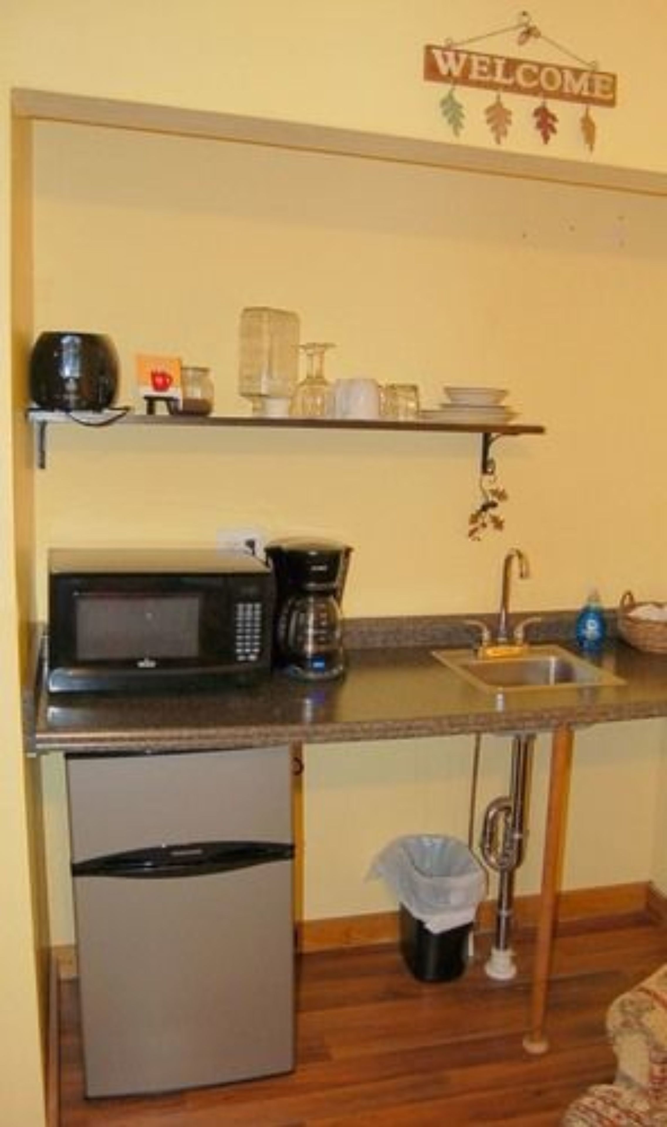 A compact kitchen with a microwave, coffee maker, and a small refrigerator. A welcome sign hangs above the counter, and a trash can is placed under the sink.
