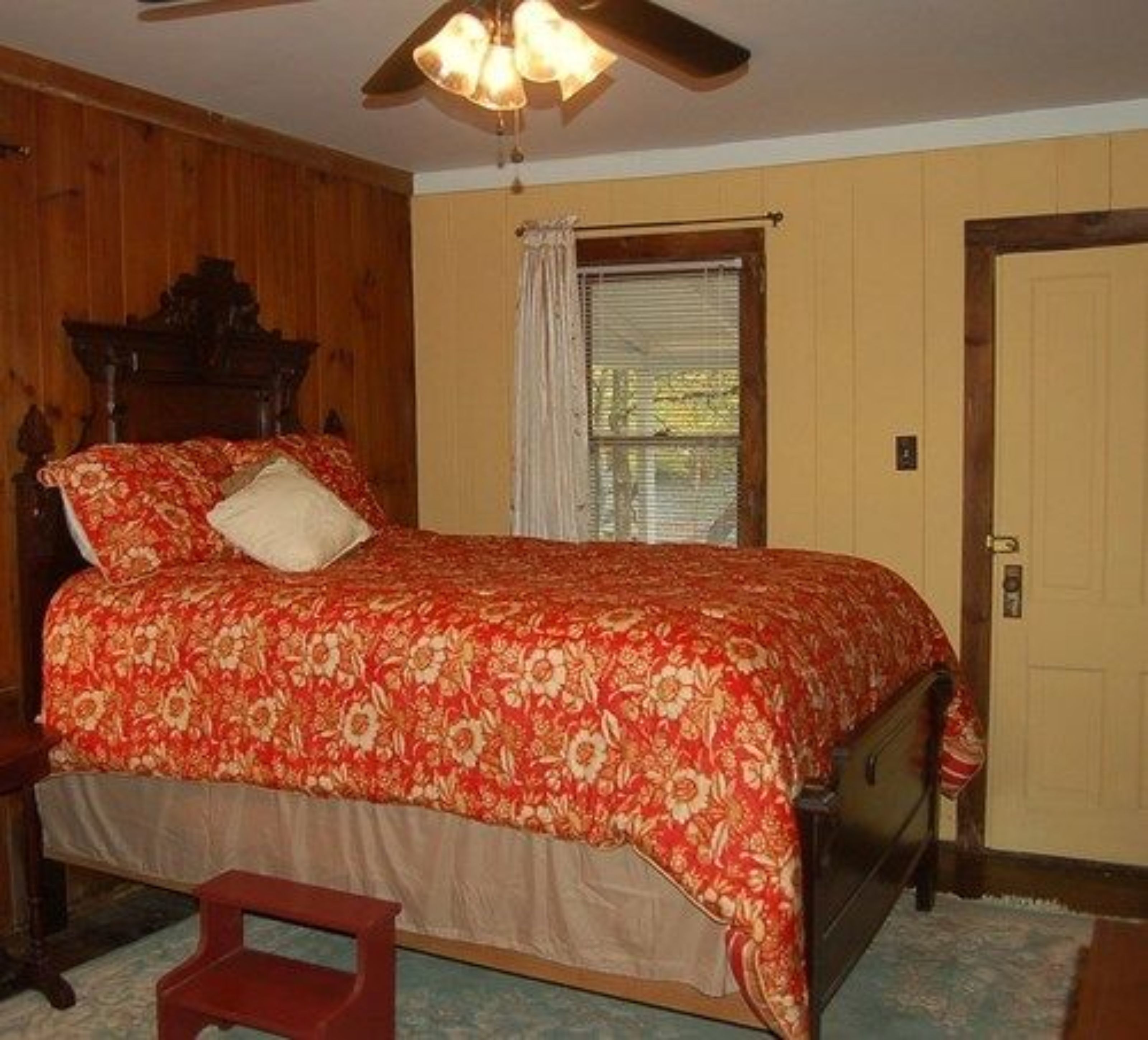 Un dormitorio con cabecera de madera, colcha estampada, cama individual, mesita de noche, una silla, ventana con persianas y una puerta.