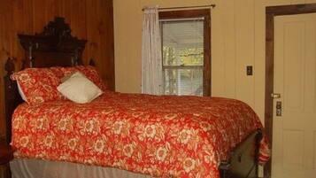 Un dormitorio con cabecera de madera, colcha estampada, cama individual, mesita de noche, una silla, ventana con persianas y una puerta.