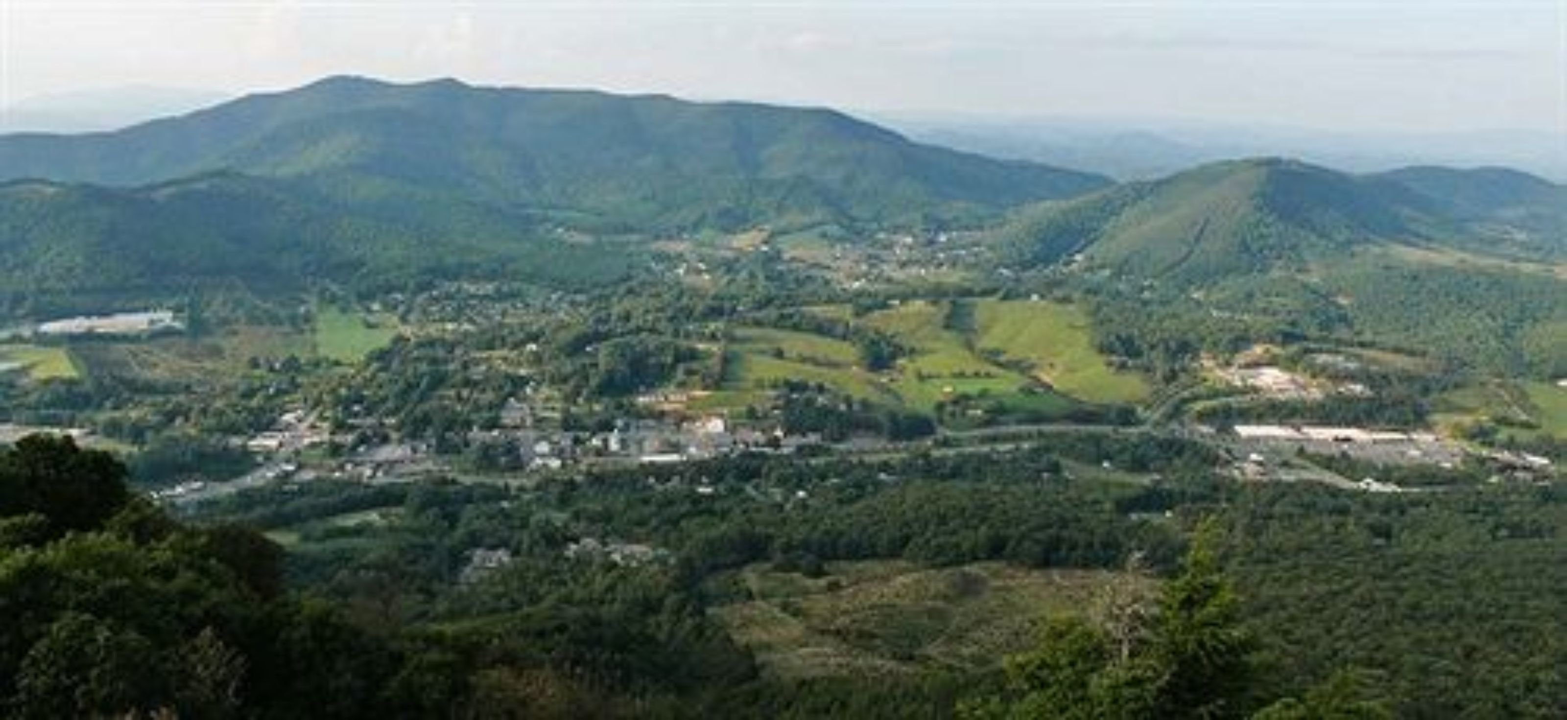 A panoramic view of a valley with scattered buildings, surrounded by hills and dense greenery.