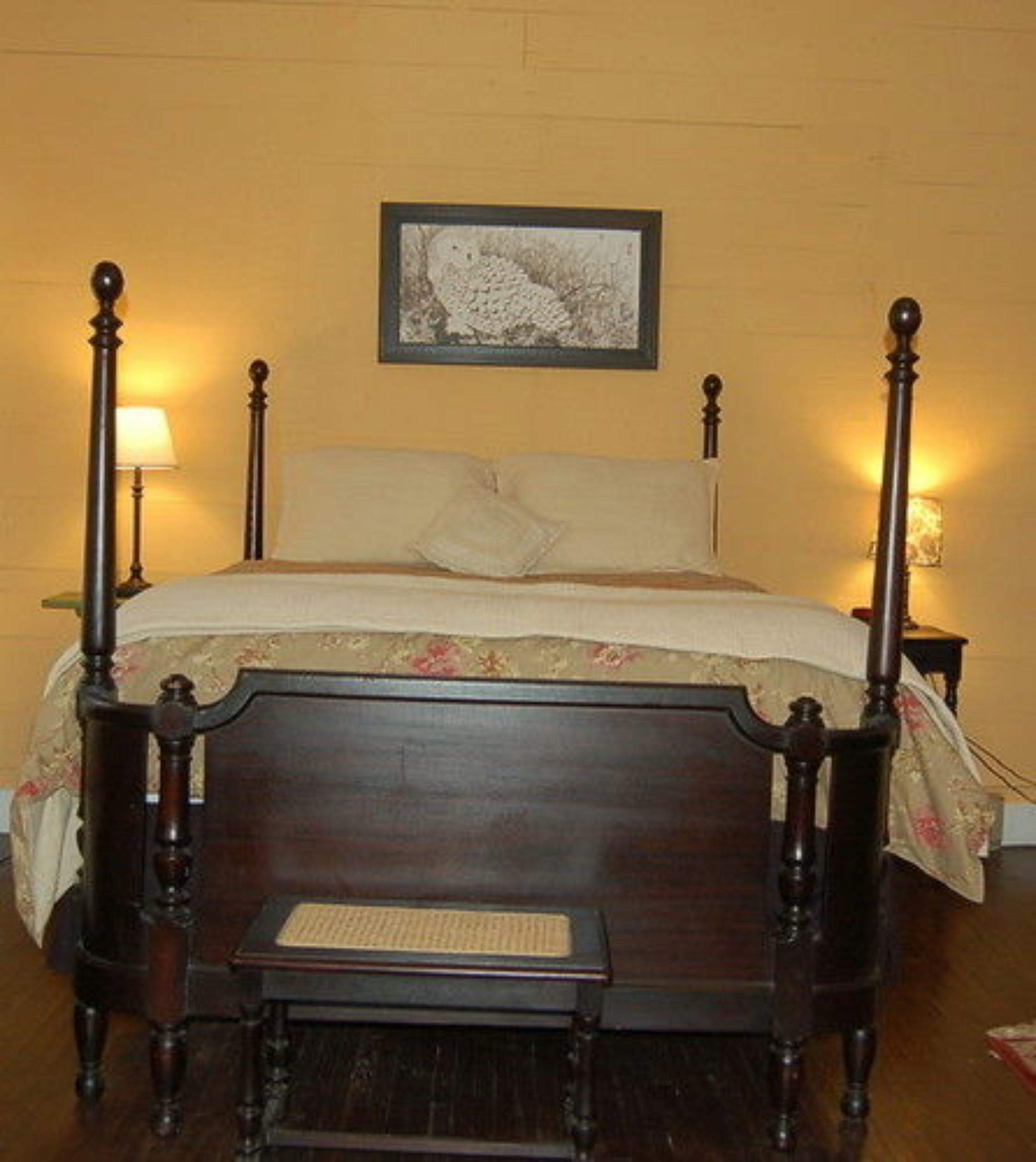 A four-poster bed with a wooden headboard and footboard, a small bedside table, and a nightstand with a lamp.