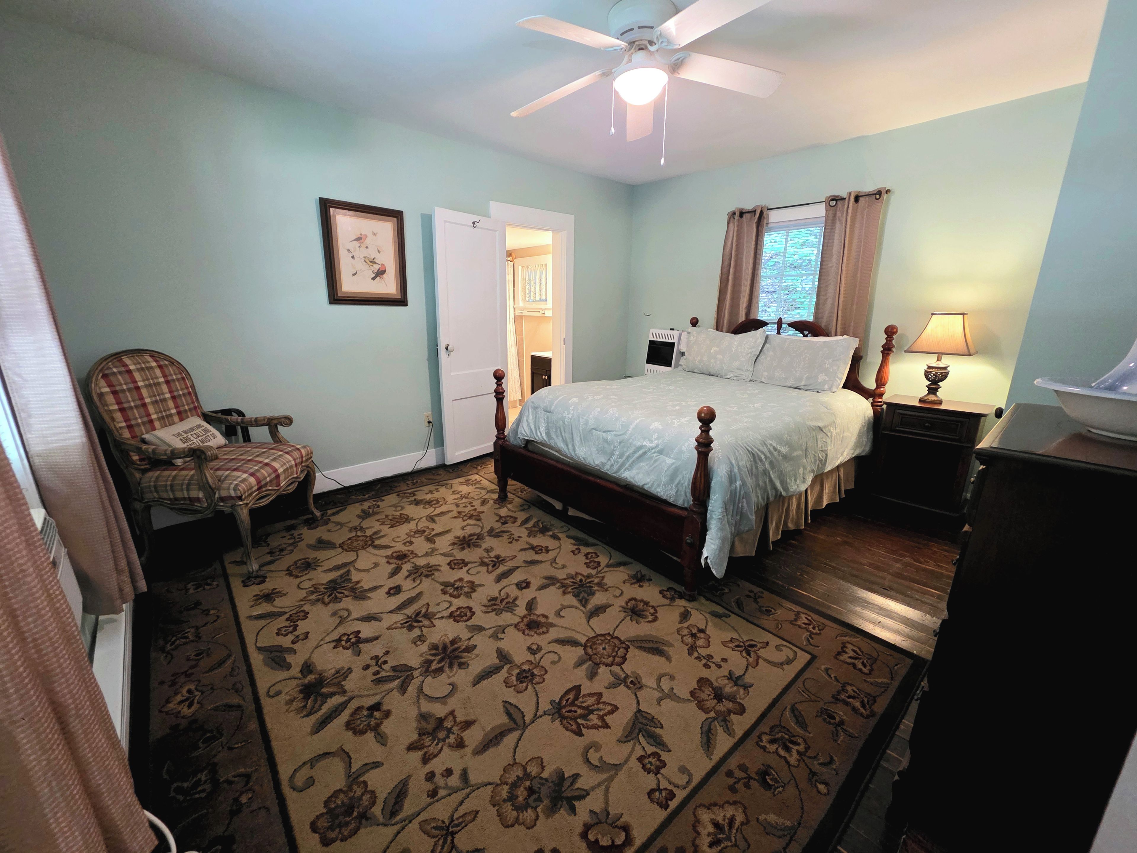 A bedroom with a bed, a chair, a nightstand, a lamp, a ceiling fan, and a door leading to another room.